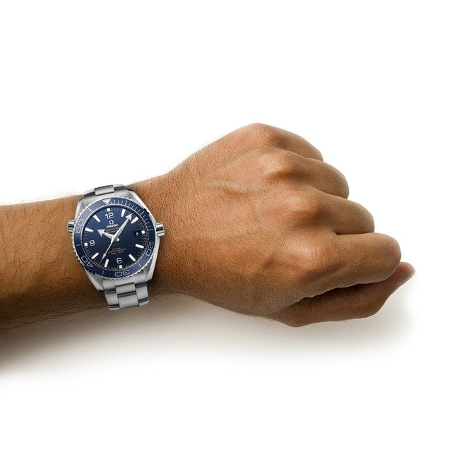 A person’s wrist wearing an OMEGA Seamaster Planet Ocean 600M, a silver metal dive watch with a blue face, on a white background.