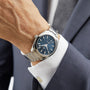 A person dressed in a navy suit and white shirt wears an OMEGA Seamaster Aqua Terra 150M, featuring a stainless steel case and blue patterned dial with silver markers.