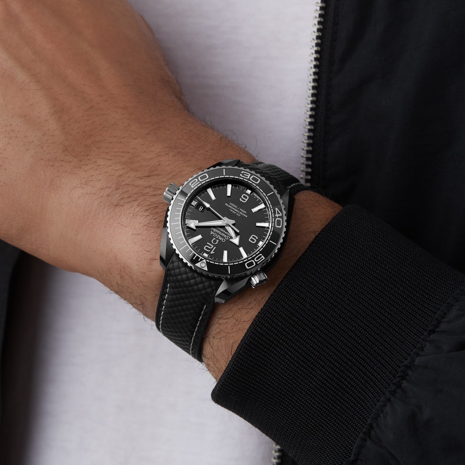 An OMEGA Seamaster Planet Ocean 600M with a textured black strap worn on the wrist, partially visible alongside a jacket and white shirt.