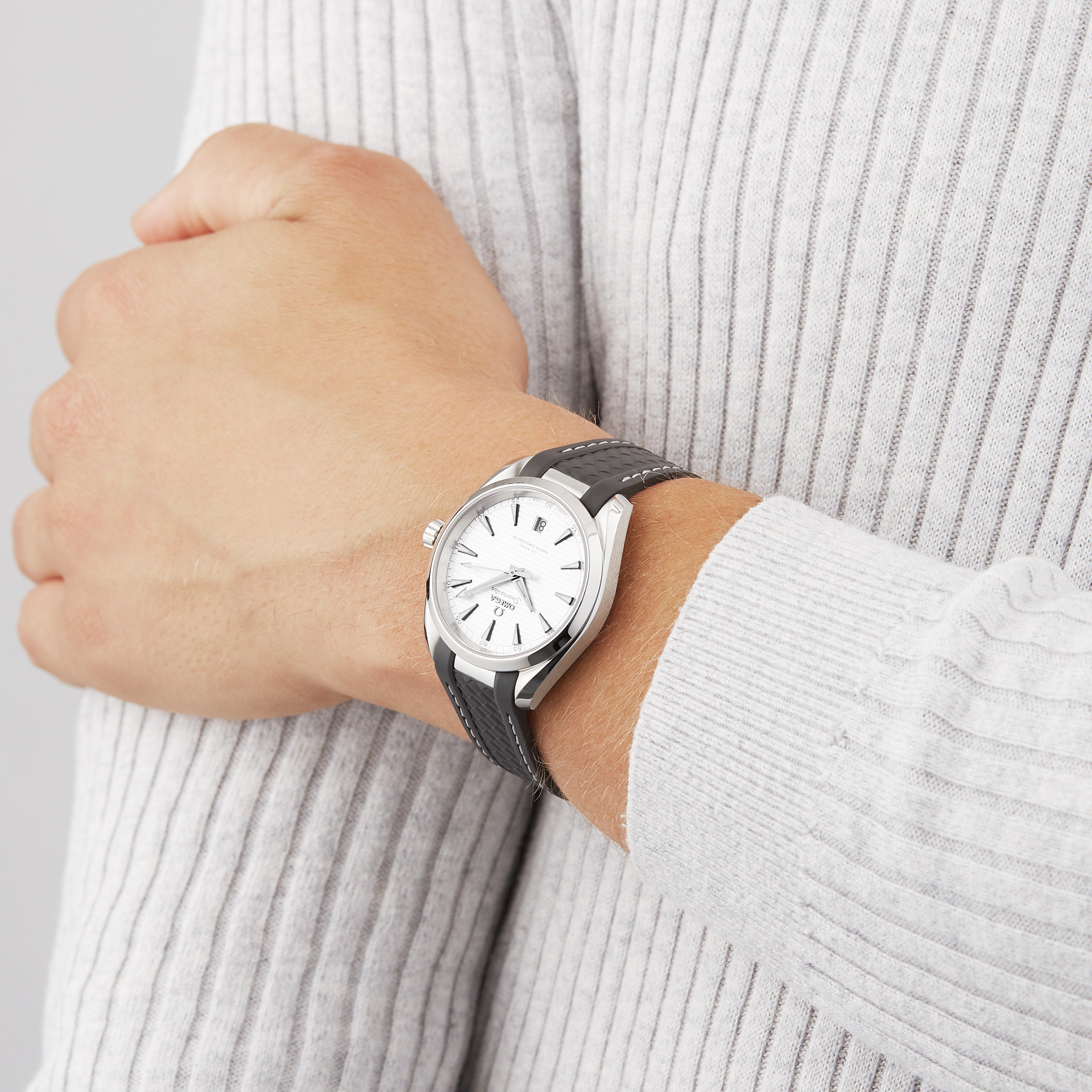 A person wearing an OMEGA Seamaster Aqua Terra 150M wristwatch with a black strap and white dial, dressed in a light gray ribbed sweater.