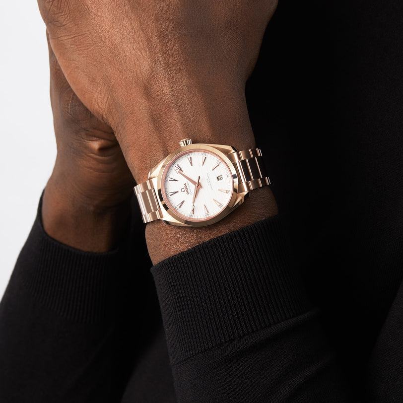 An individual in a black long-sleeve top wears an OMEGA Seamaster Aqua Terra 150M with 18K Sedna™ gold accents, a white dial, and a metal bracelet on their left wrist.