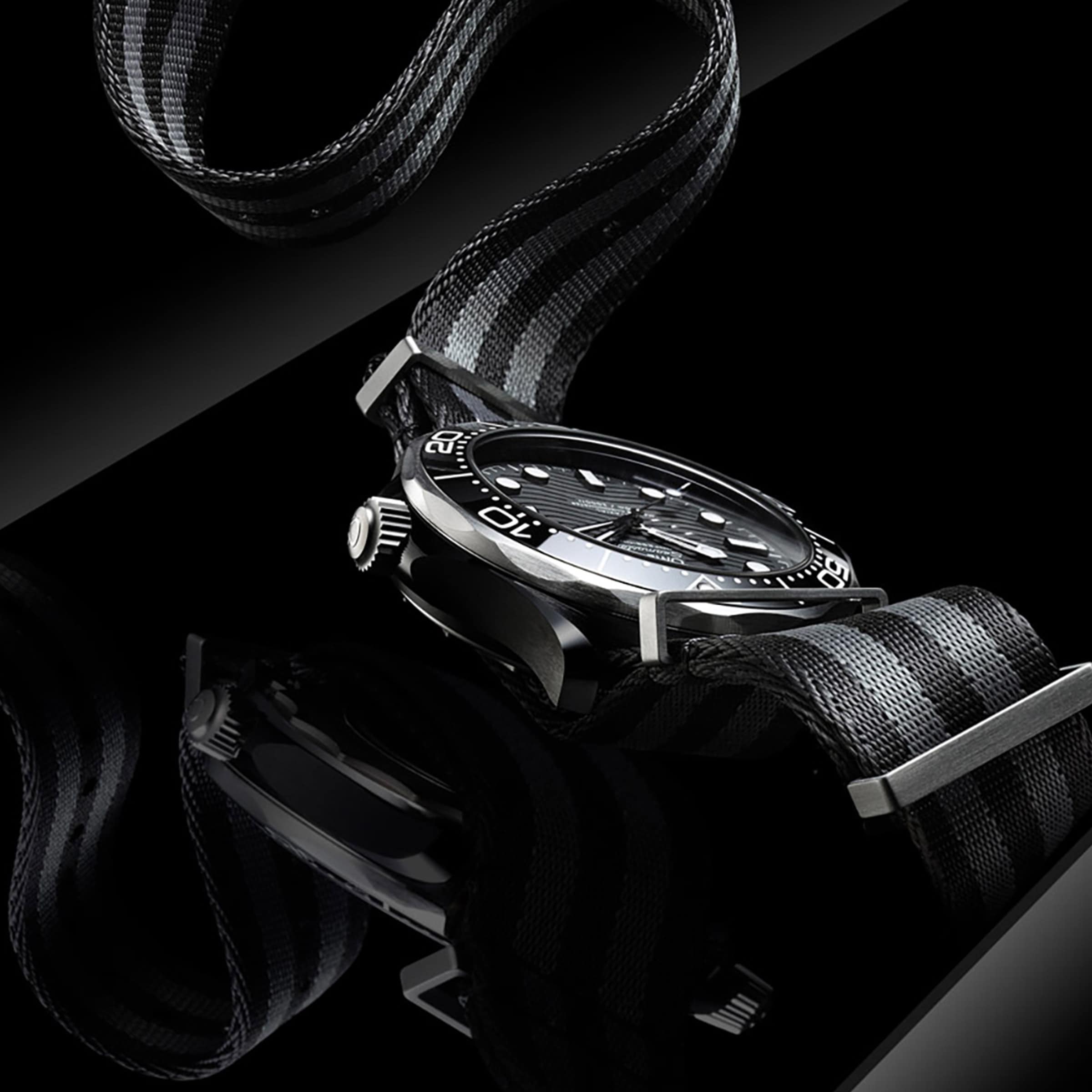 A close-up of the OMEGA Seamaster Diver 300M, a black and silver dive watch with a black and gray striped nylon strap, displayed on a reflective black surface.