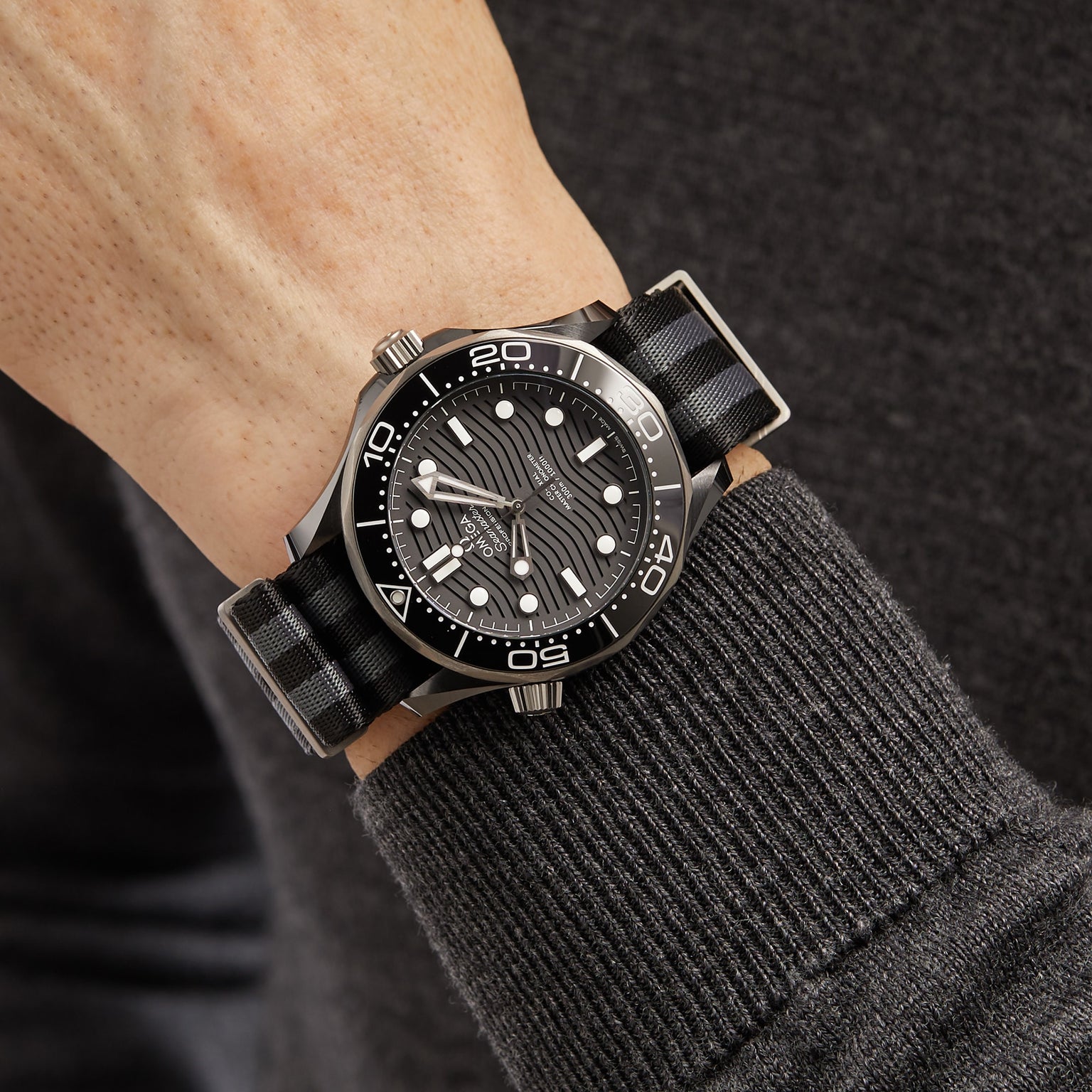 A person wearing an OMEGA Seamaster Diver 300M with a black dial, white markers, stainless steel case, and a black and gray striped strap over a dark long sleeve shirt.