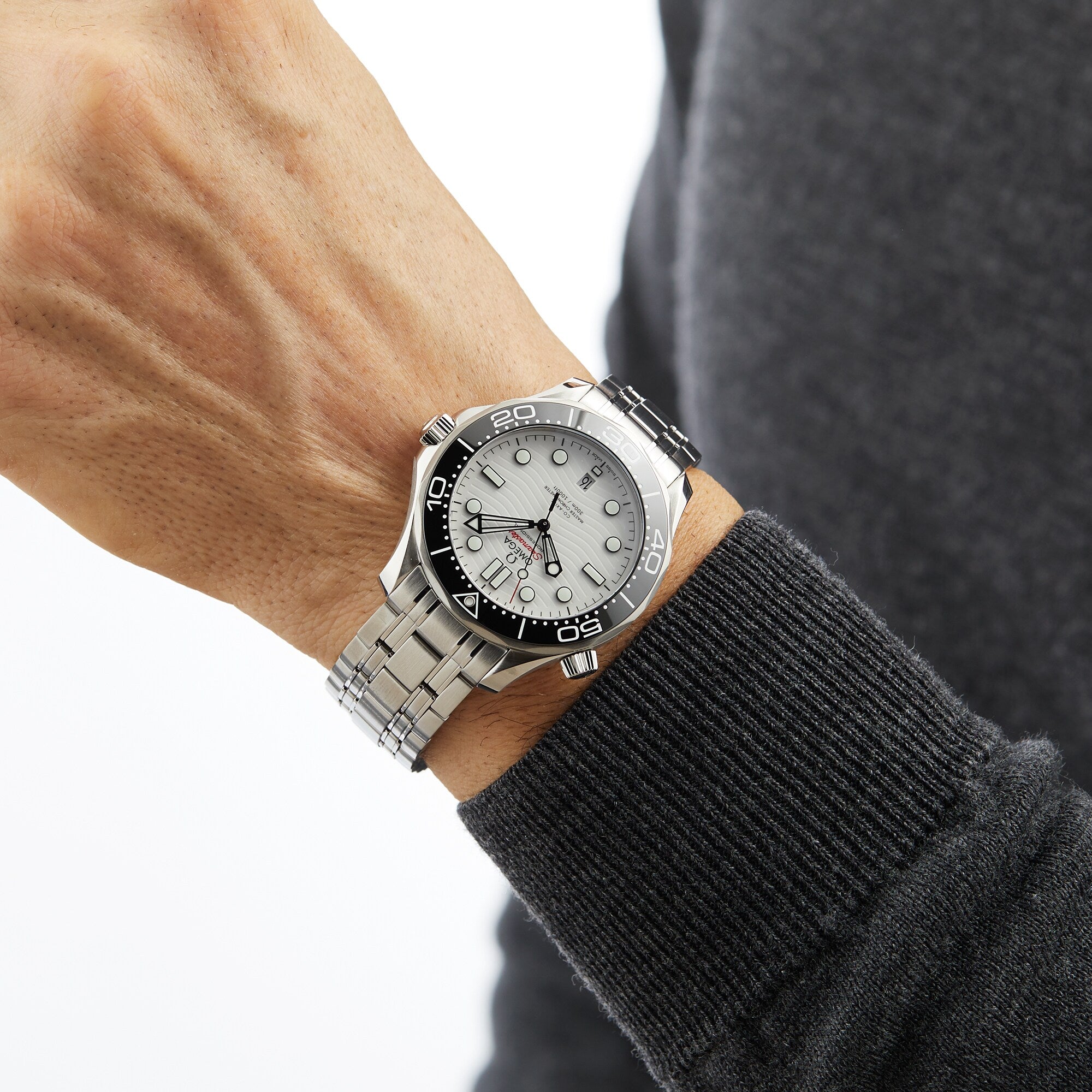 A person wears an OMEGA Seamaster Diver 300M with a black bezel and silver dial, featuring the OMEGA Calibre 8800, on their left wrist against a gray long sleeve shirt.