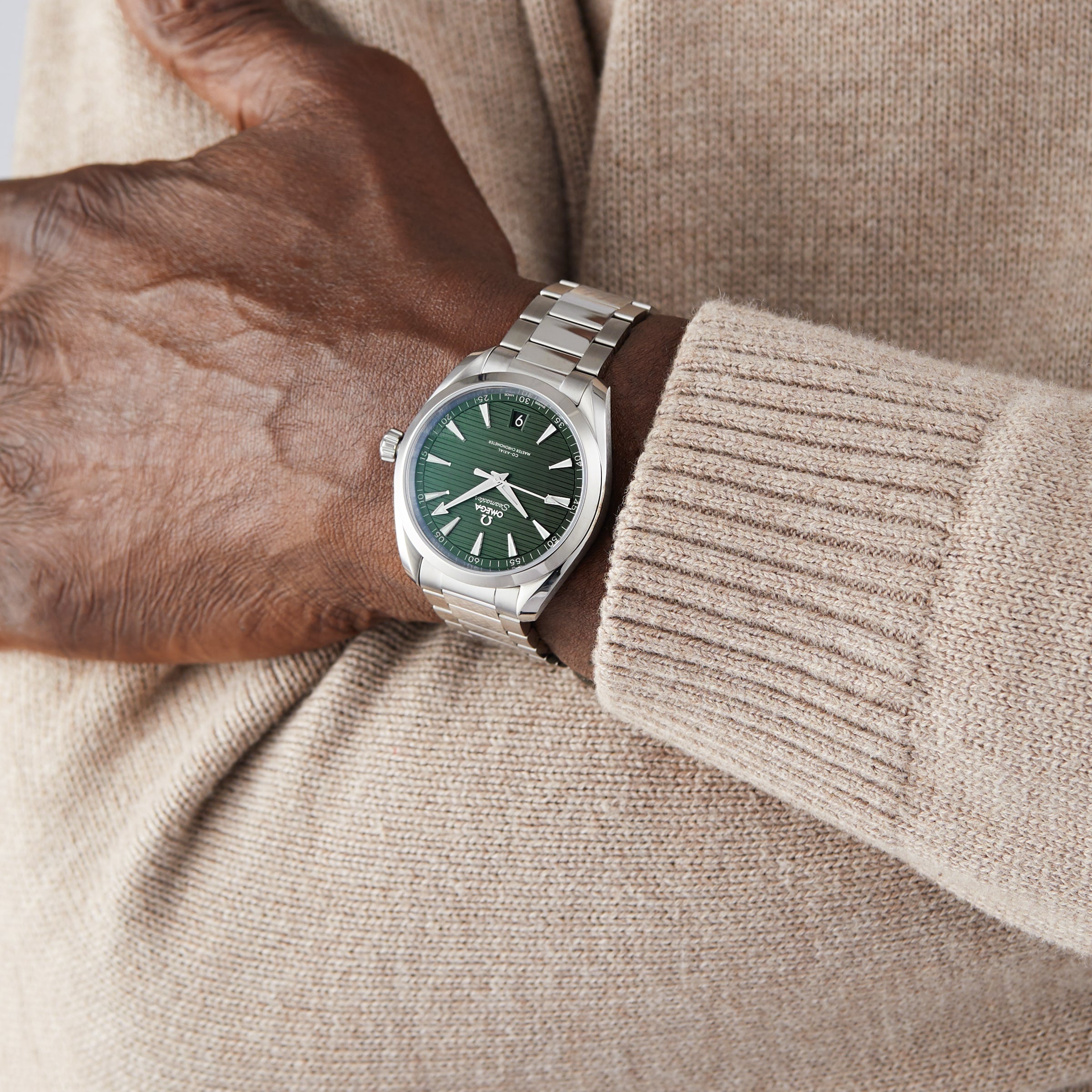 A person in a beige sweater wears an OMEGA Seamaster Aqua Terra 150M with a green dial, featuring the Master Chronometer 8900 calibre, and crosses their arm over their chest.