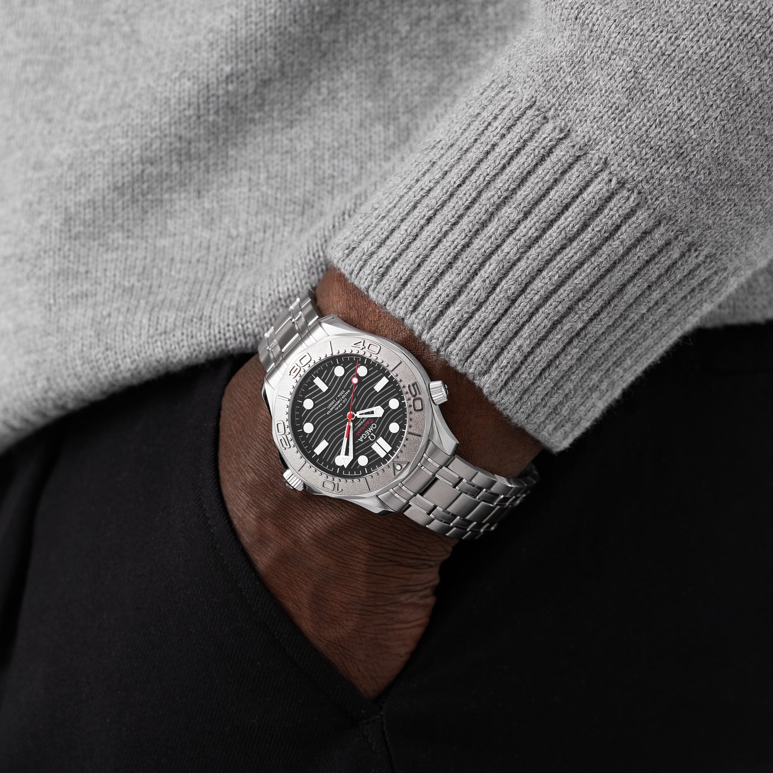 A person in a gray sweater and black pants has their hand in their pocket, showcasing the OMEGA Seamaster Diver 300M Nekton Edition, featuring a laser-ablated ceramic dial and stainless steel case.
