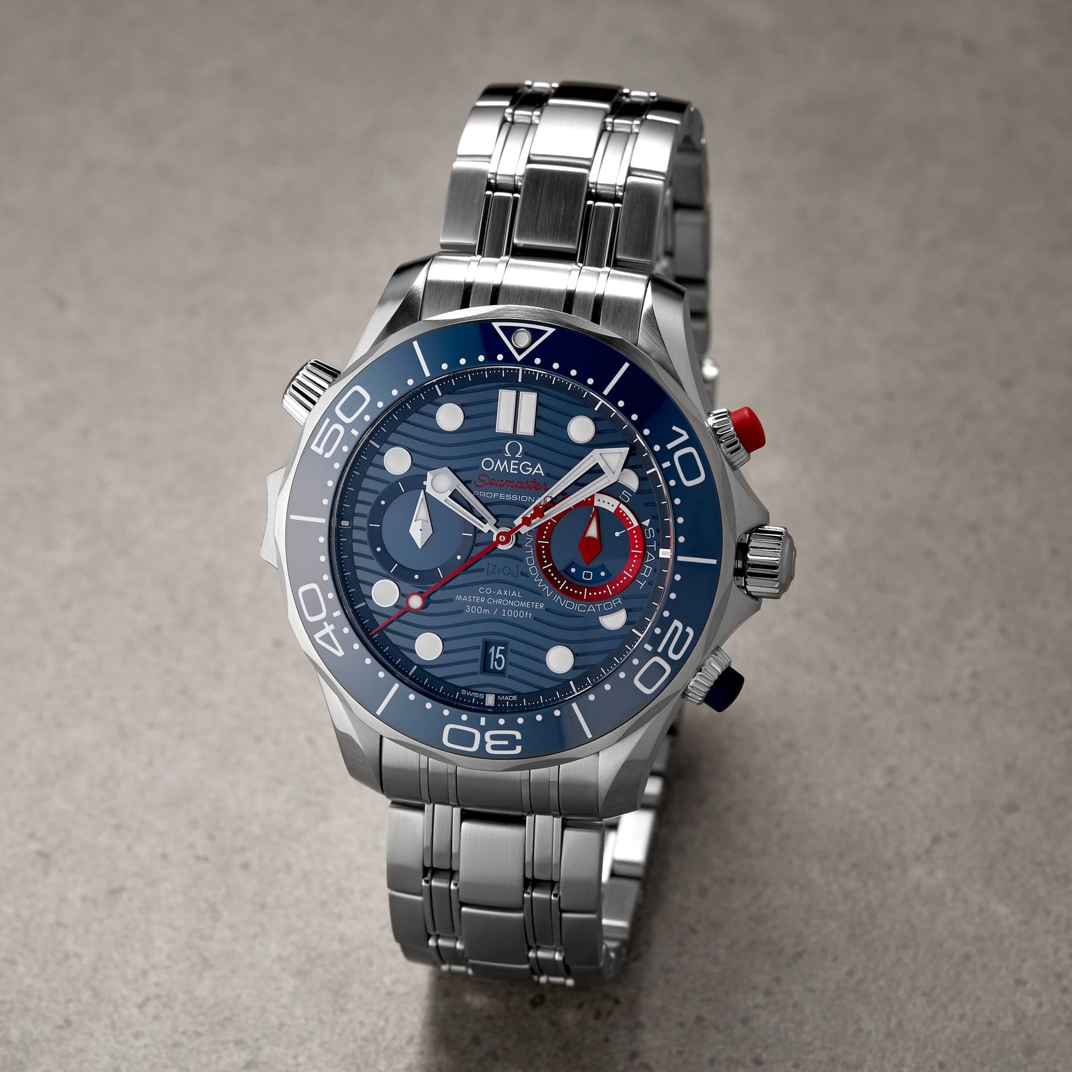 An OMEGA Seamaster Diver 300M Americas Cup Edition wristwatch with a blue dial, white markers, red and blue accents, and a metal bracelet is displayed on a grey surface.