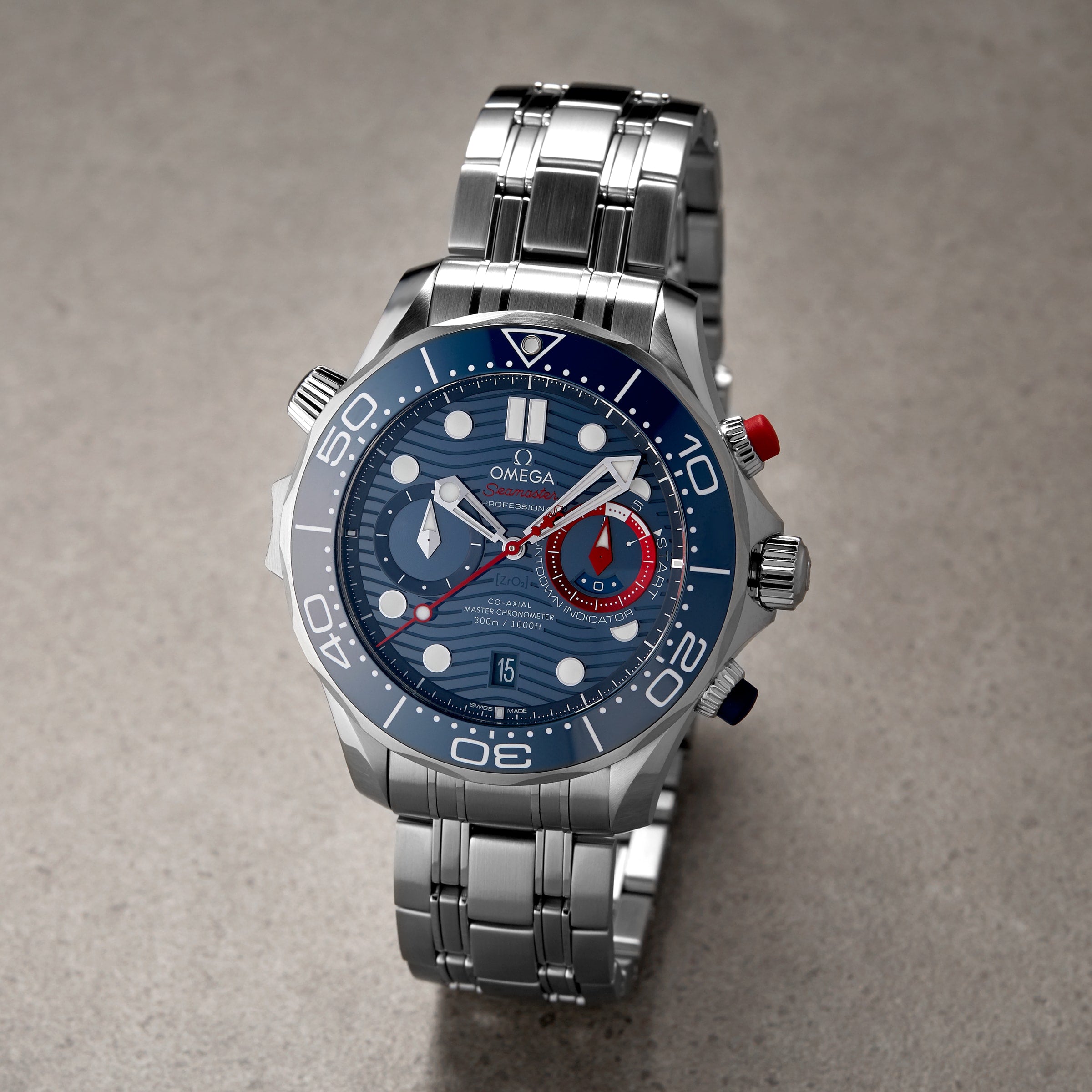 An OMEGA Seamaster Diver 300M Americas Cup Edition wristwatch with a blue dial, white markers, red and blue accents, and a metal bracelet is displayed on a grey surface.
