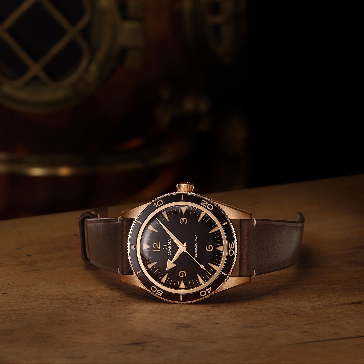 An OMEGA Seamaster 300 wristwatch with a gold case and brown leather strap rests on a wooden surface, with a brass diving helmet in the background.