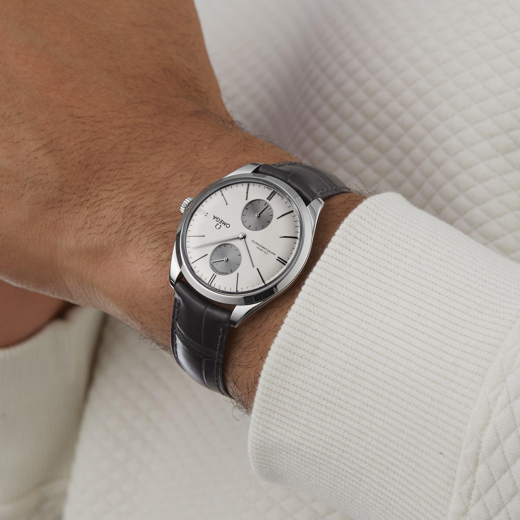An individual wears an OMEGA De Ville Trésor stainless steel analog wristwatch with a leather strap, paired with a white textured long-sleeve shirt.