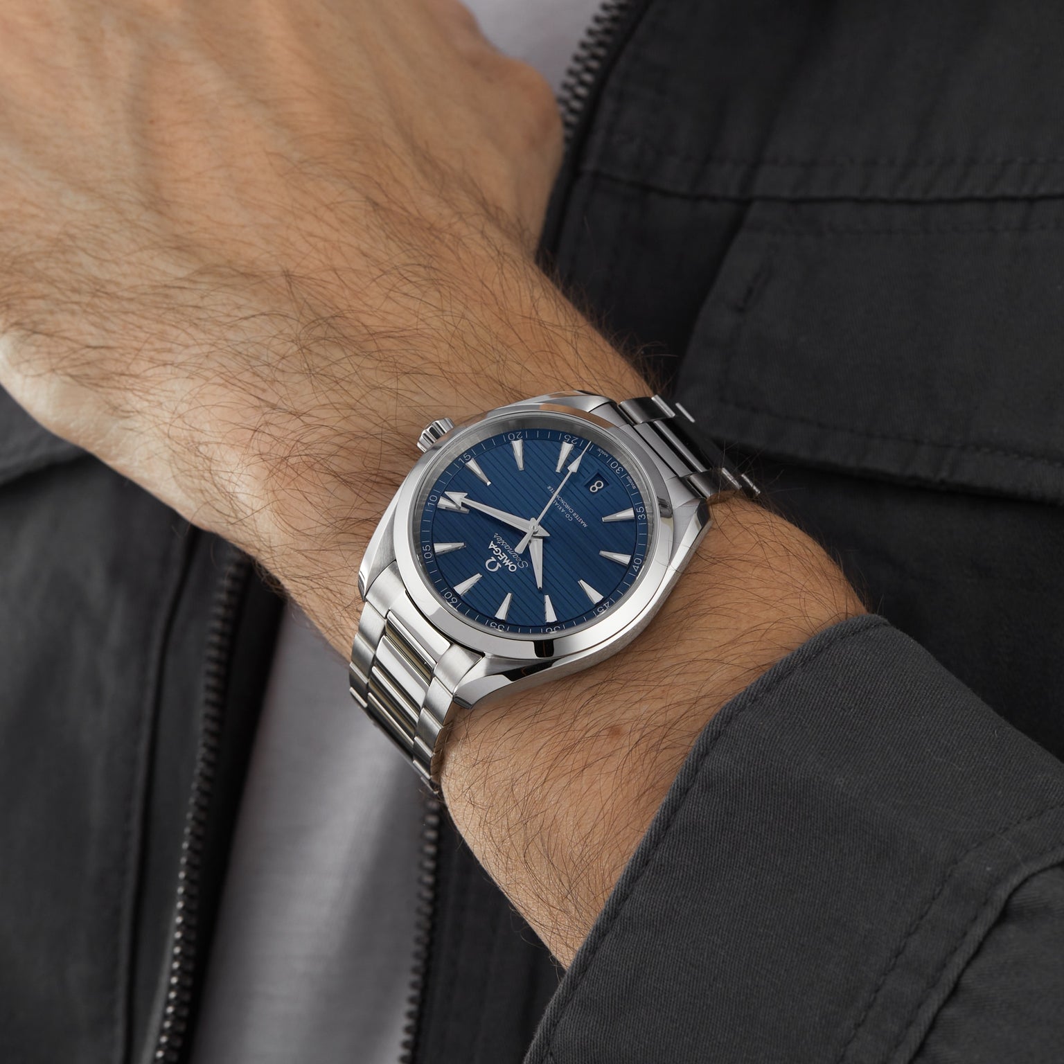 A person wearing an OMEGA Seamaster Aqua Terra 150M with a blue face and metal band is photographed from the side against a dark jacket, highlighting the watch’s maritime heritage.