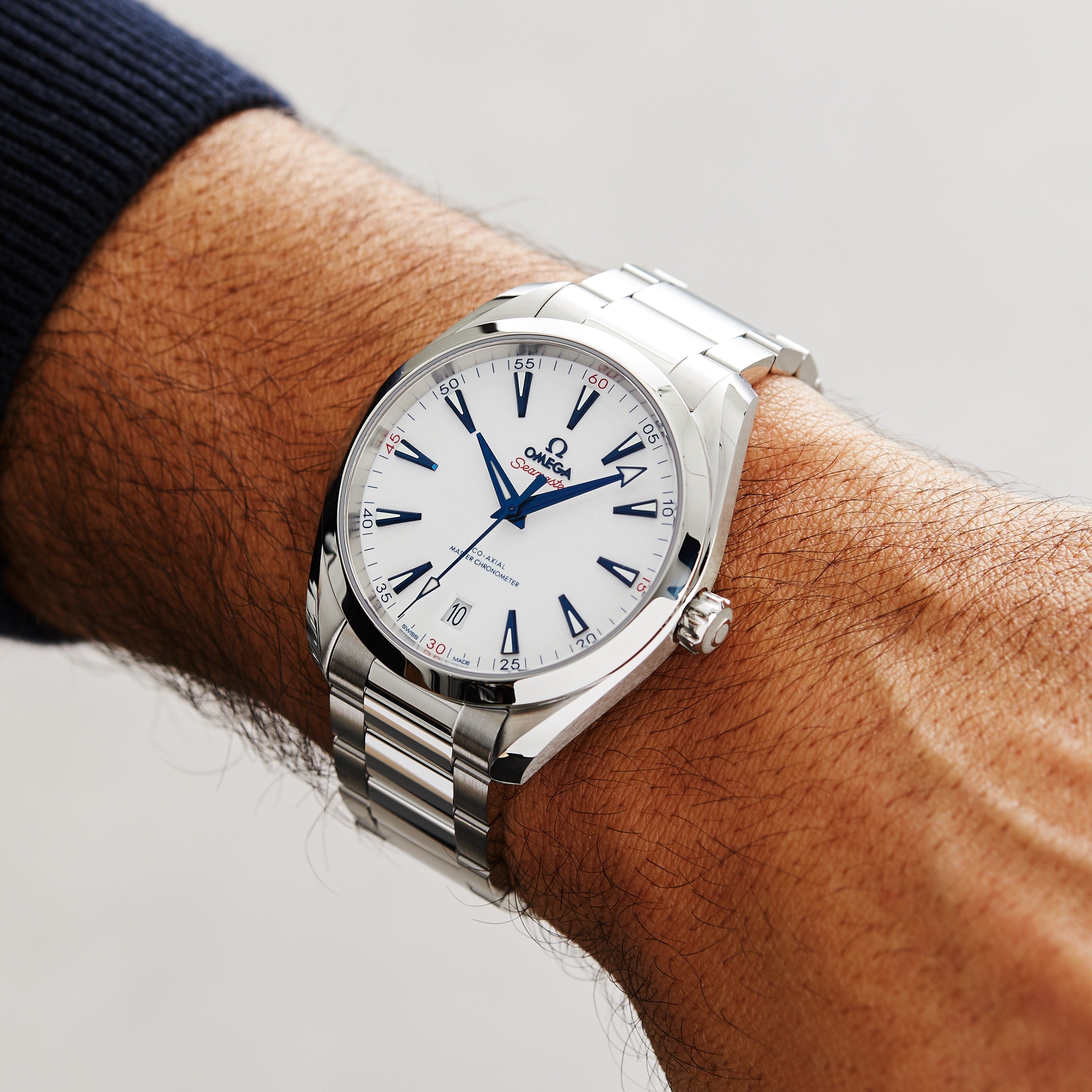 A person wears an OMEGA Seamaster Aqua Terra 150M with a silver case, white dial, blue hour markers, and 8900 calibre on their left wrist. This elegant watch celebrates the Olympic Winter Games 2022.