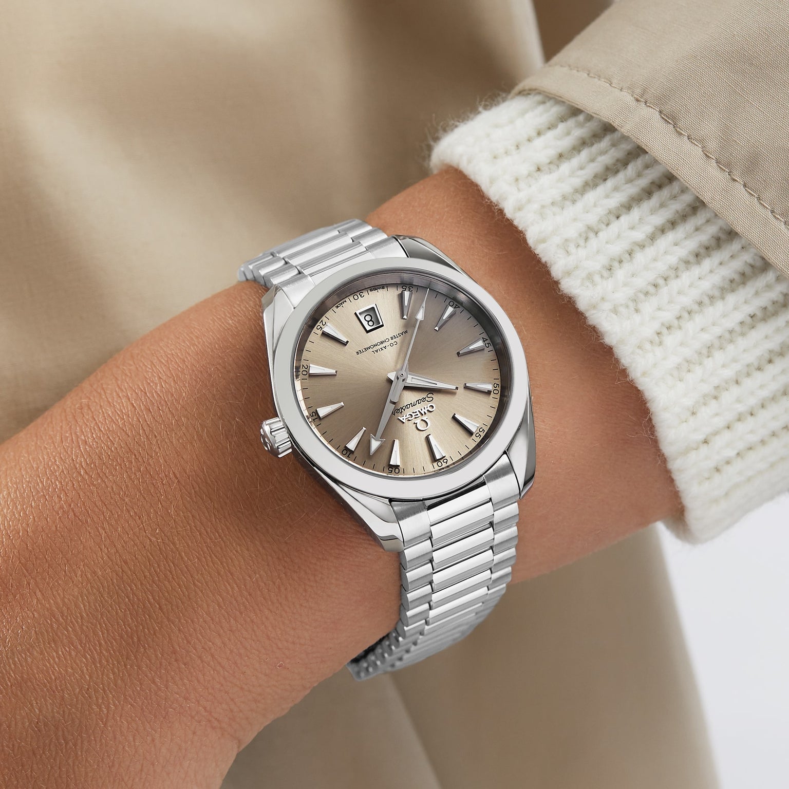 A person wearing an OMEGA Seamaster Aqua Terra 150M with a sandstone dial and stainless steel bracelet, styled with a light knit sweater and beige coat.