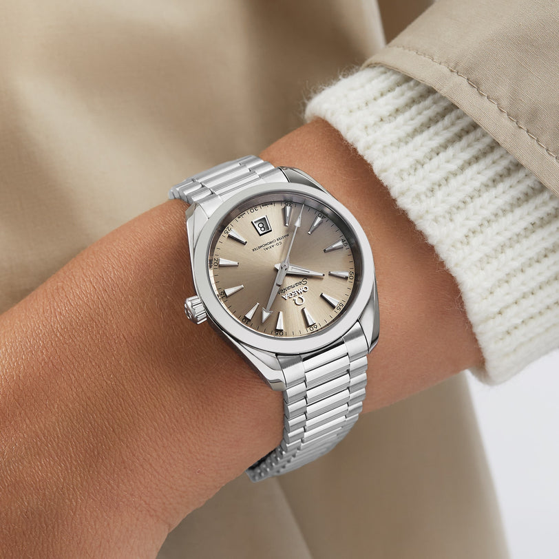 A person wearing an OMEGA Seamaster Aqua Terra 150M with a sandstone dial and stainless steel bracelet, styled with a light knit sweater and beige coat.