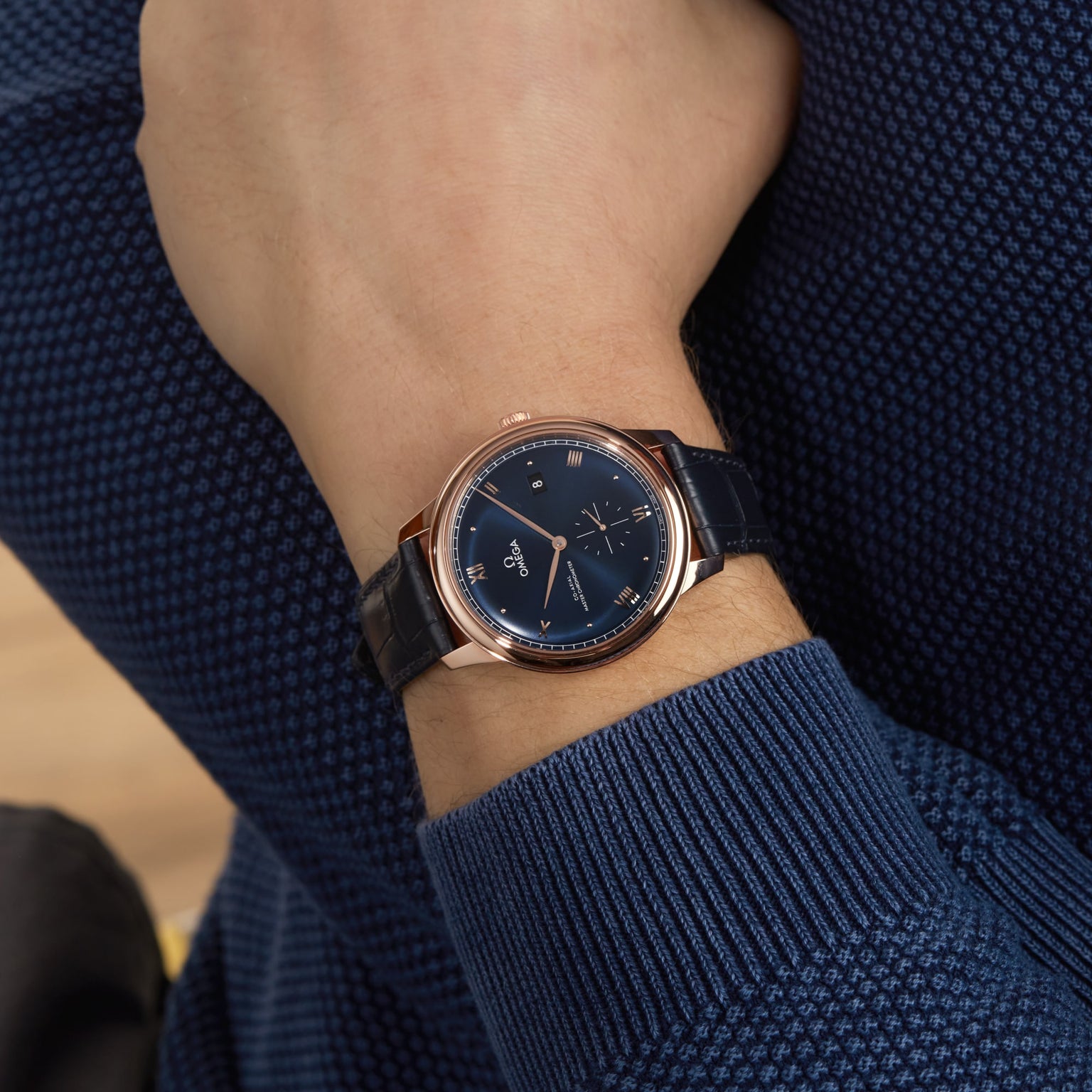 A person wearing a blue long-sleeve shirt and an OMEGA De Ville Prestige wristwatch with a blue dial, black leather strap, and Master Co-Axial 8802 movement.