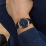 A person wearing a blue long-sleeve shirt and an OMEGA De Ville Prestige wristwatch with a blue dial, black leather strap, and Master Co-Axial 8802 movement.