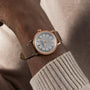 A close-up of a wrist in a beige sweater wearing the Raymond Weil Millesime Automatic Silver Sector Dial, a Swiss-made gold watch with a white face and beige leather strap.