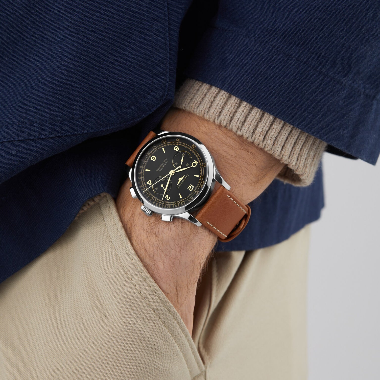 Wearing a Longines Record Chronograph with a brown leather strap, black dial, and stainless steel case, the person pairs it with a blue jacket, beige sweater, and light khaki pants, hand casually in their pocket.