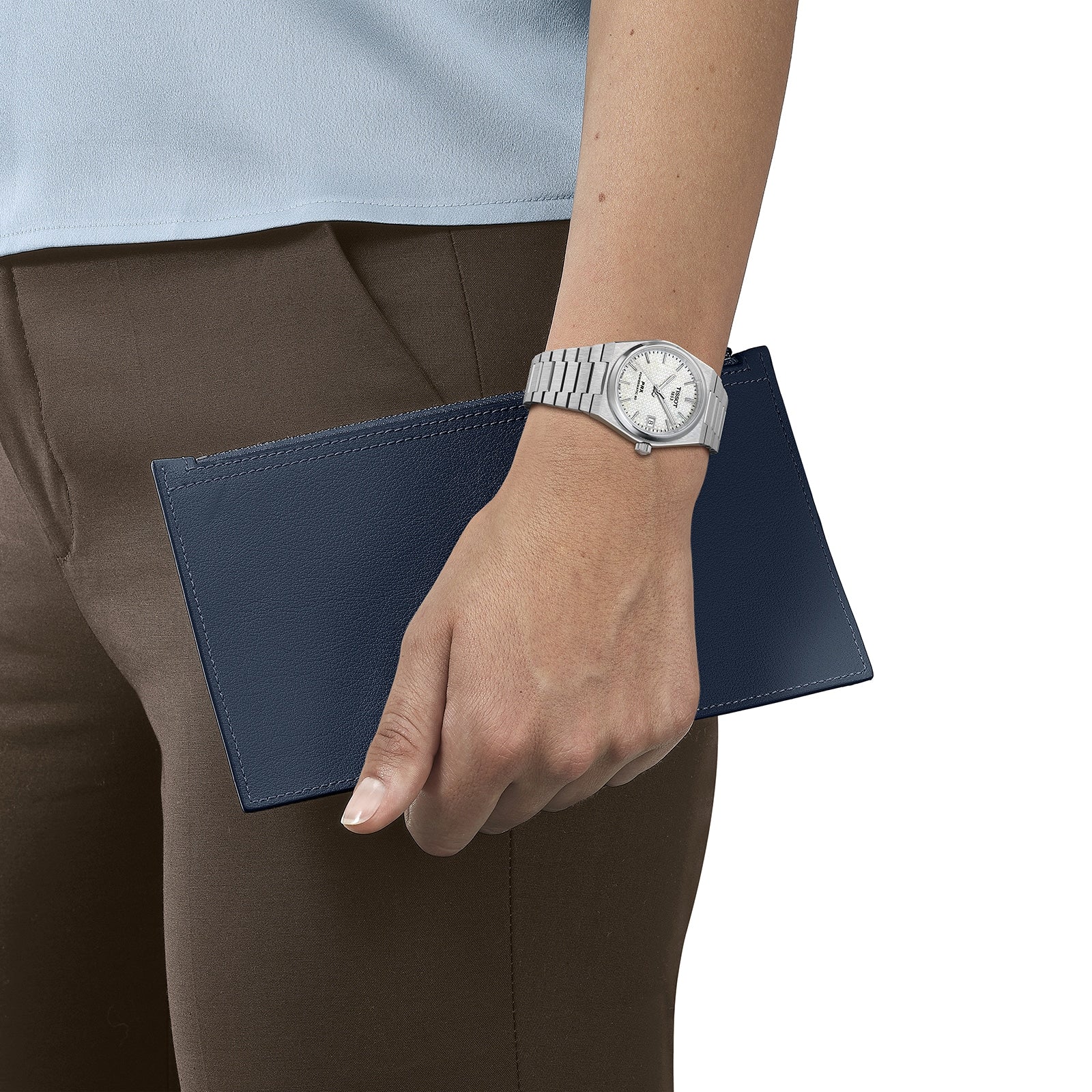 A person wearing a Tissot PRX Powermatic 80 holds a dark blue wallet or clutch against their brown pants and light blue top.