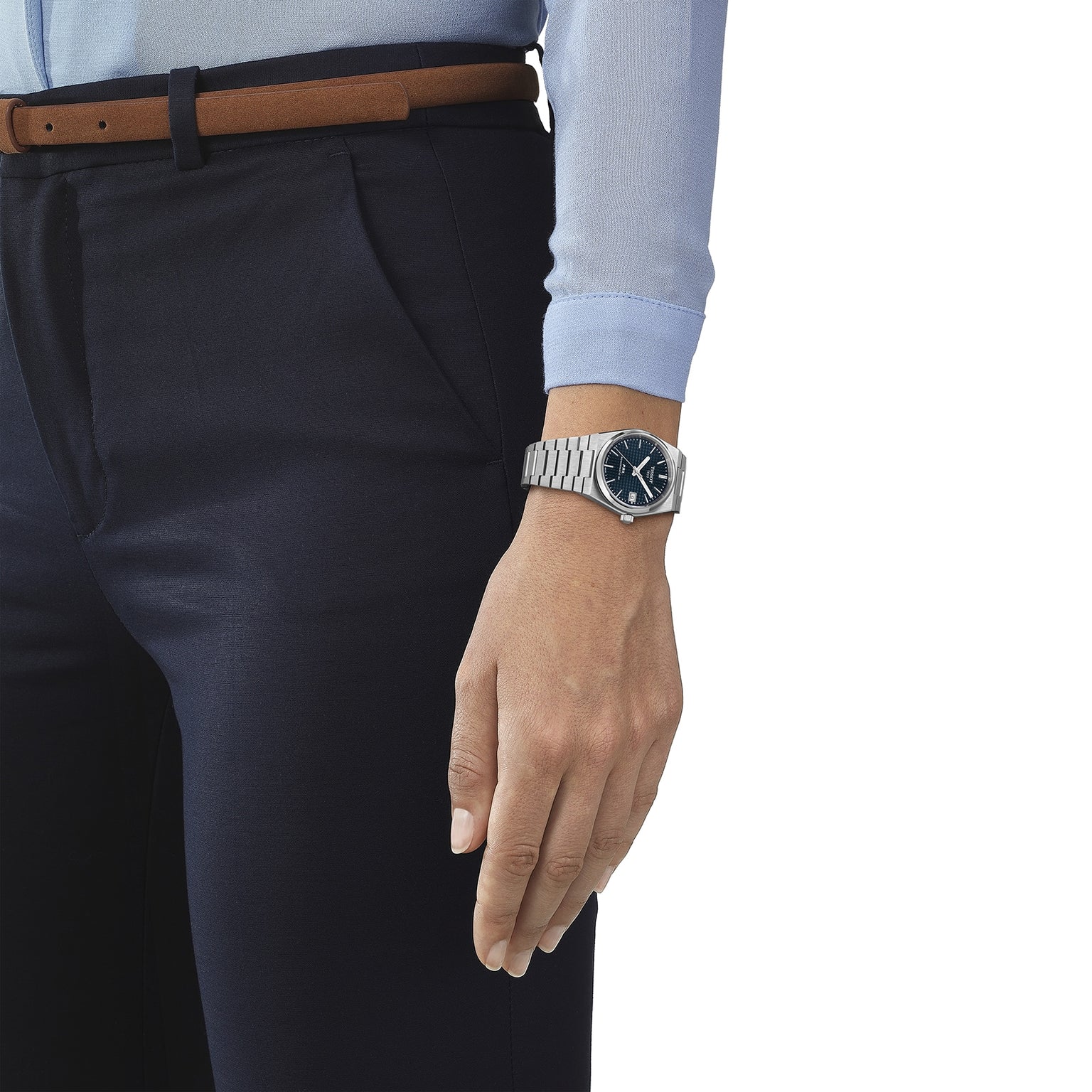 A person in dark trousers and a light blue shirt stands with their arm relaxed at their side, wearing a Tissot PRX Powermatic 80 with a striking blue dial.