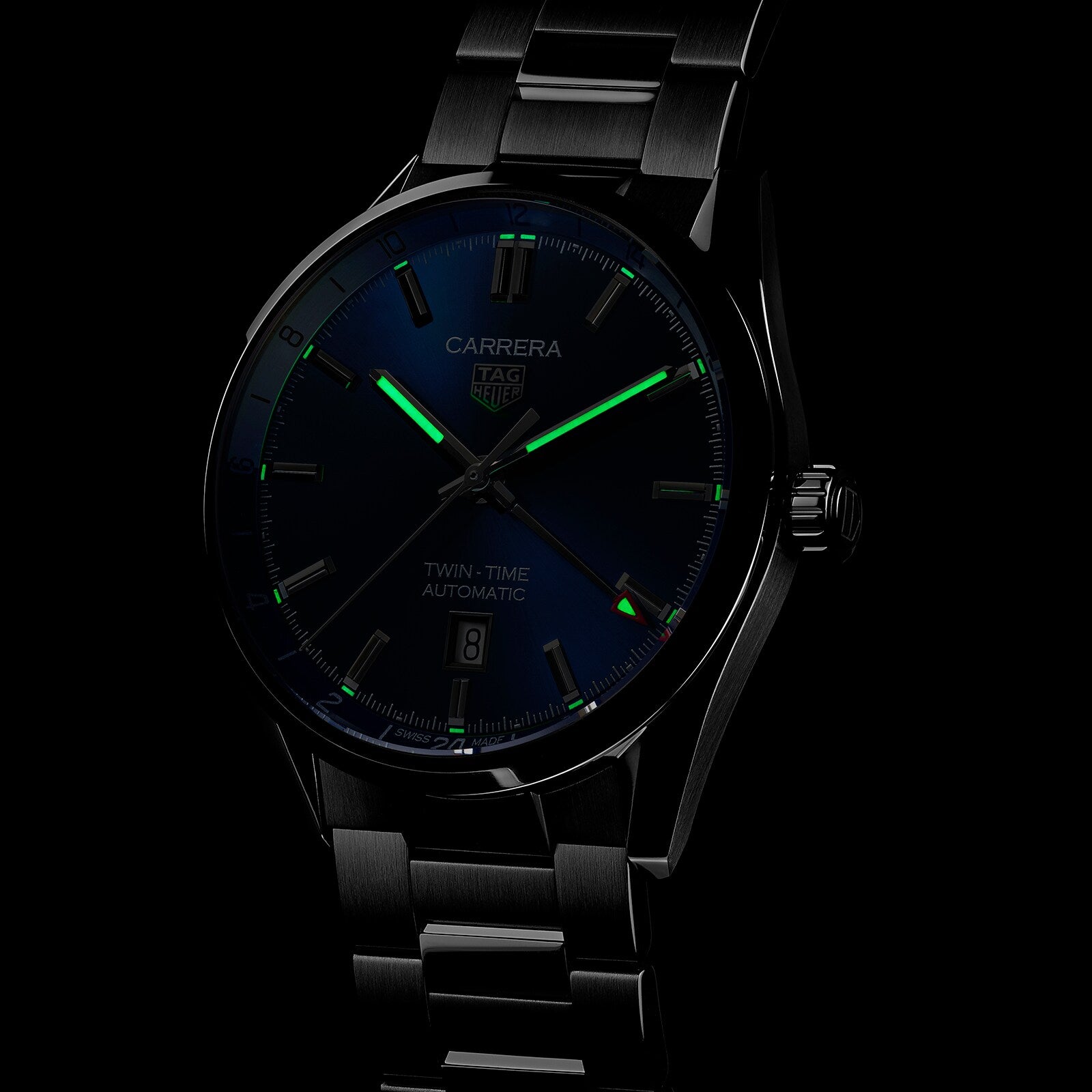 The TAG Heuer Carrera Twin-Time watch features GMT functionality, a dark dial with luminous green hands, and a stainless steel bracelet, shown against a black background.
