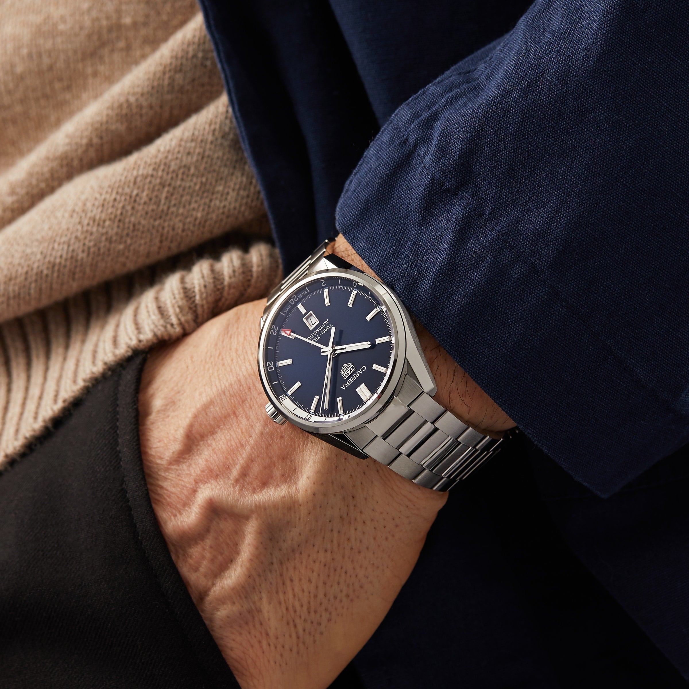 A person wearing a TAG Heuer Carrera Twin-Time silver wristwatch with a blue face and metal band, featuring GMT function, dressed in layers: beige sweater, black shirt, and blue jacket.