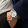Wearing a TAG Heuer Carrera Date silver wristwatch with a white dial and metal band, the person has their hand in the pocket of dark pants, dressed in a grey shirt and navy jacket.