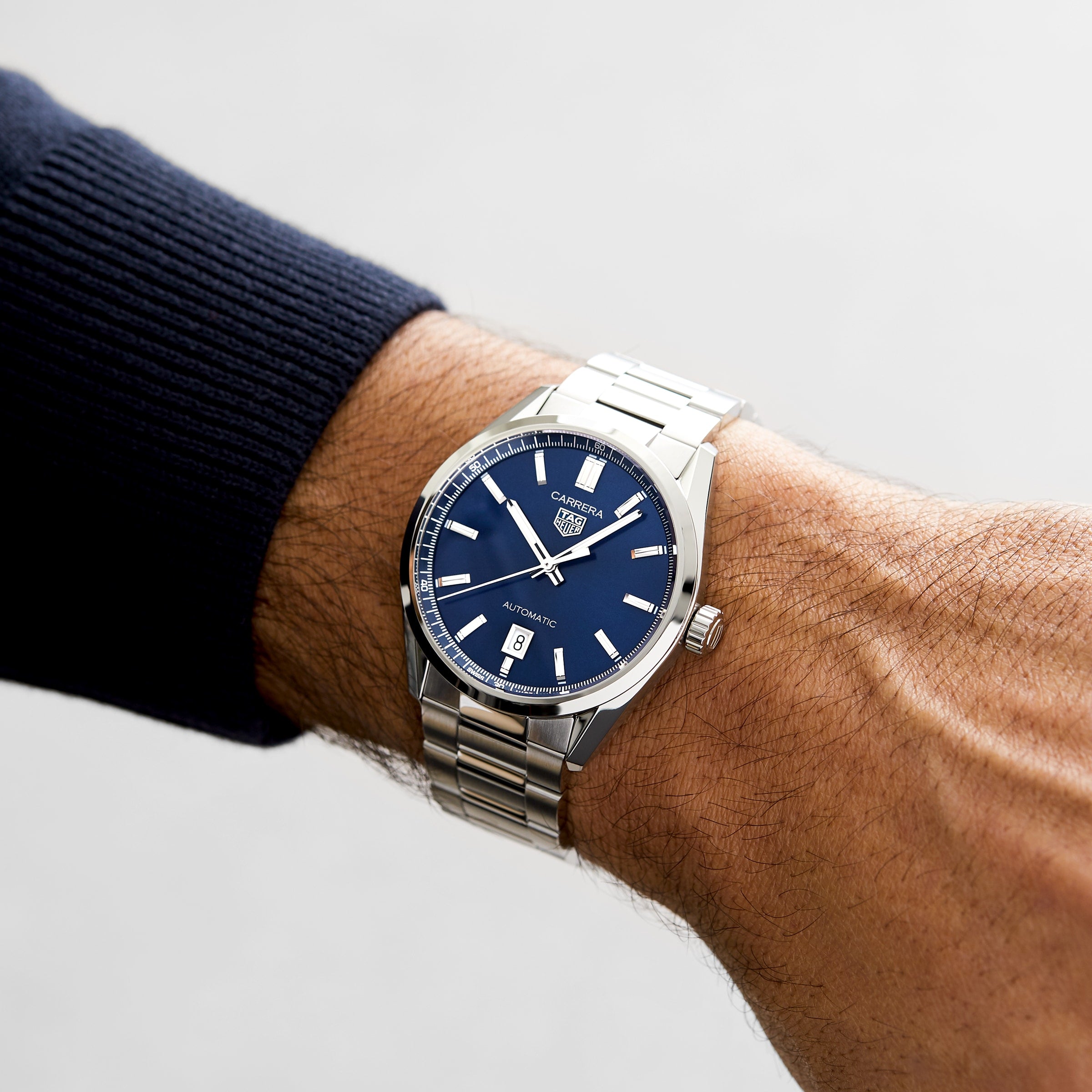 A person wearing a TAG Heuer Carrera Date watch with a stainless steel case, blue dial, date display, and automatic movement on their left wrist.
