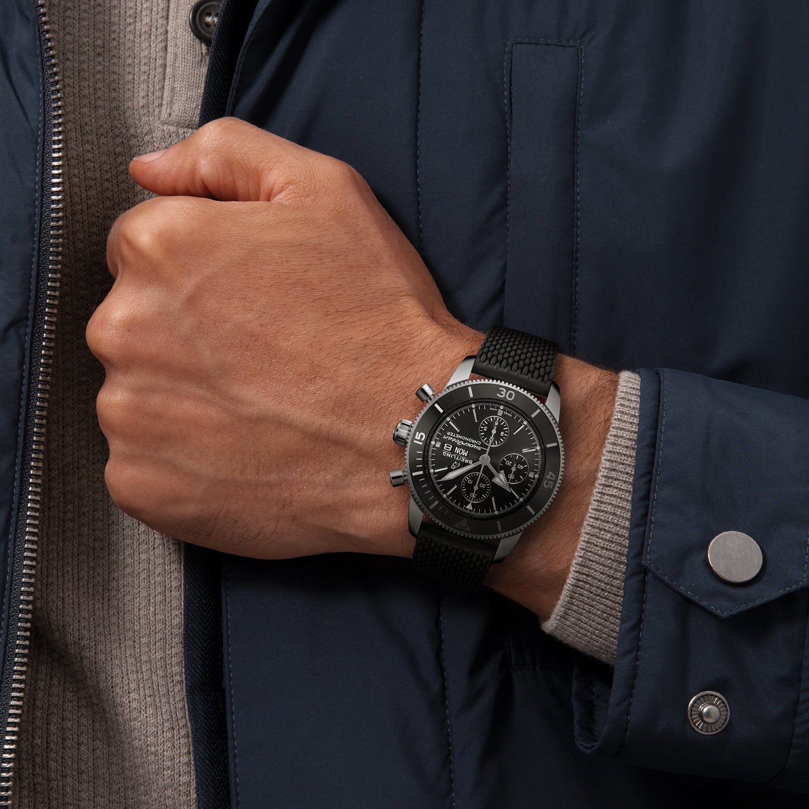 A person wearing a Breitling Superocean Heritage Chronograph 44 with a black strap, dressed in a navy blue jacket and beige sweater.