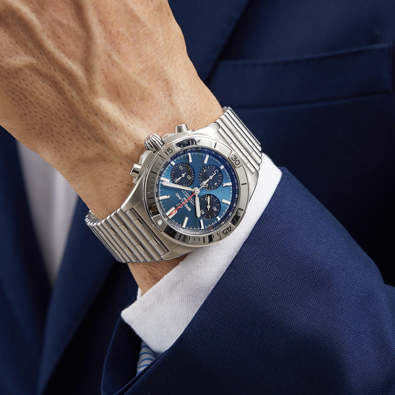 A person in a blue suit wears a Breitling Chronomat B01 42, a mechanical watch with a blue dial, metal bracelet, and the renowned Breitling 01 movement.