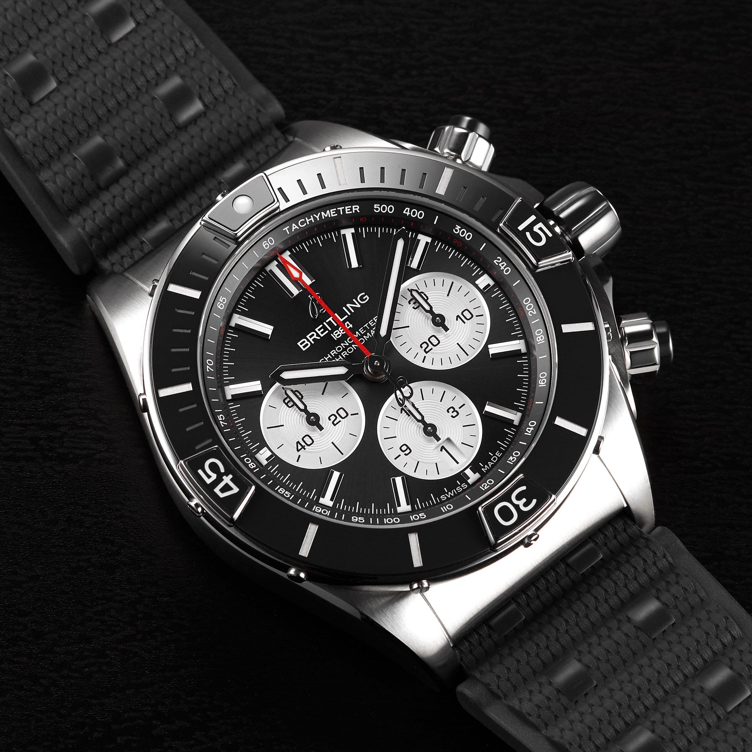The Breitling Super Chronomat B01 44 chronograph features a black bezel, three sub-dials, a tachymeter scale, and a black textured strap with the precise Breitling 01 COSC-certified calibre, elegantly displayed on a dark surface.