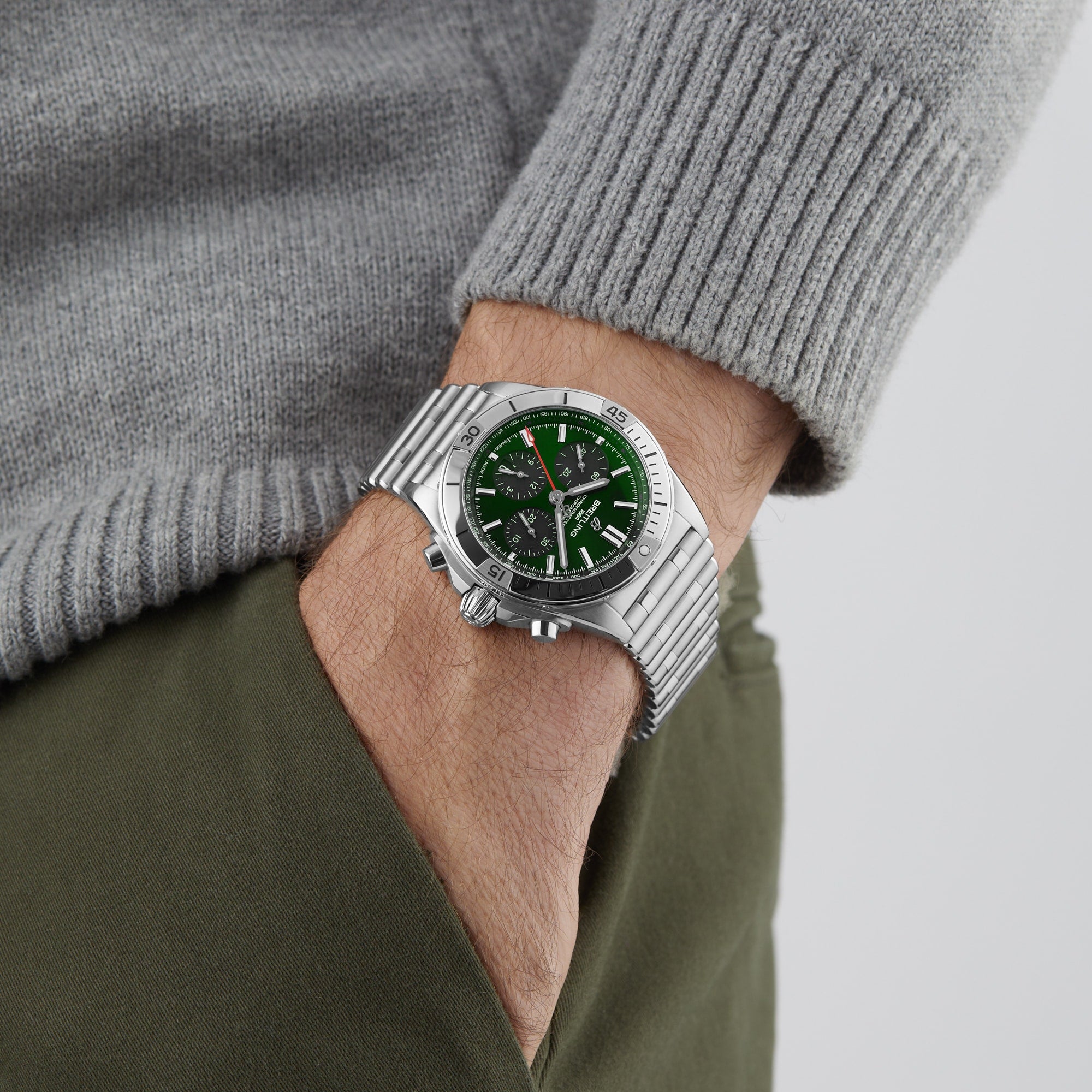 Wearing a Breitling Chronomat B01 42 with a green dial and stainless steel case, paired with a grey sweater and green pants, a person stands casually with one hand in their pocket.