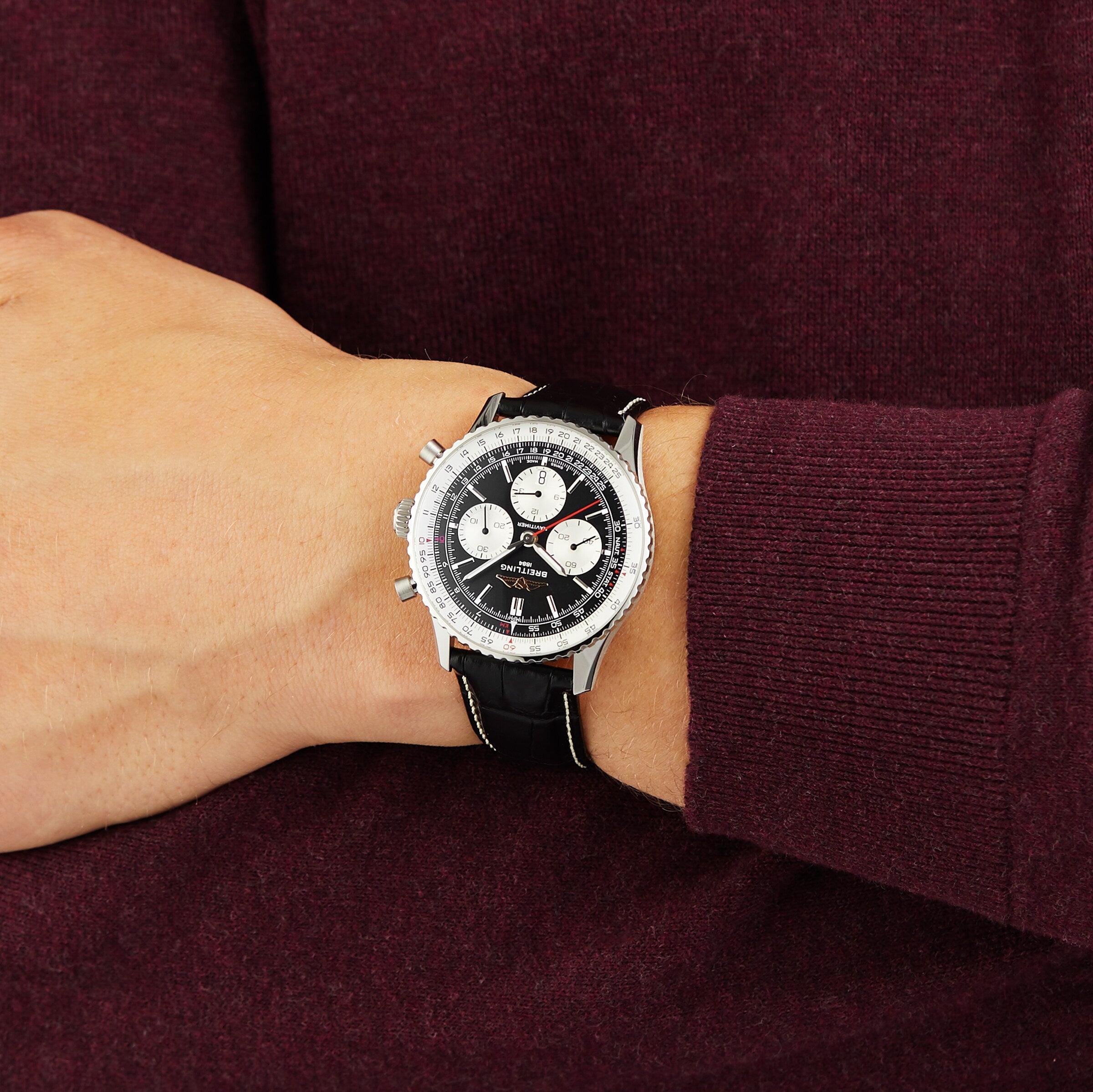 A person in a long-sleeve burgundy top wears a Breitling Navitimer B01 Chronograph 43 pilots watch, featuring a silver and black dial with a black leather strap.
