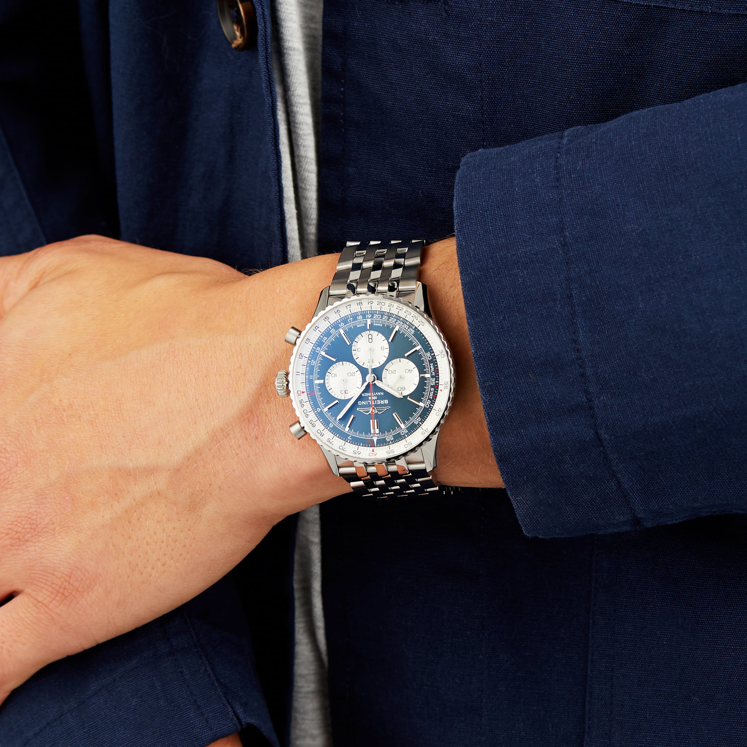 A person in a blue jacket showcases a Breitling Navitimer B01 Chronograph 46 on their left wrist, featuring a silver case, blue dial, and three subdials.