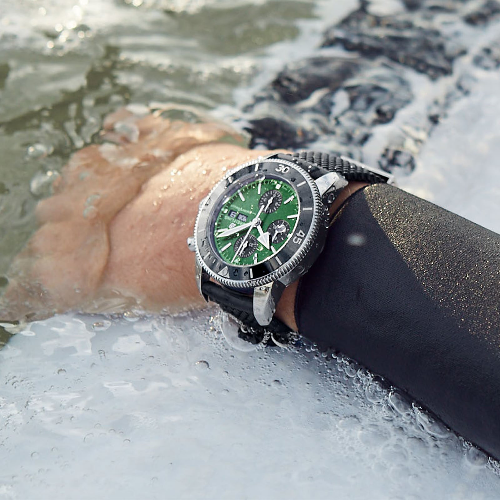 A persons wetsuit-clad arm is partially submerged in water, showcasing a large green and black Breitling Superocean Heritage Chronograph 44 on their wrist.