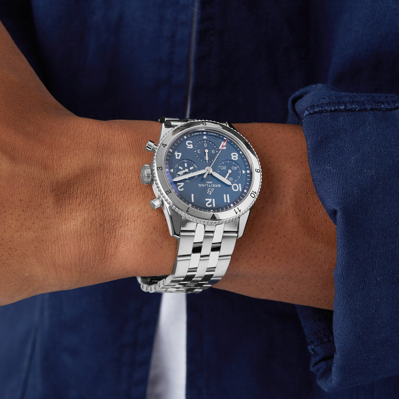 A person wearing the Breitling Classic AVI Chronograph 42 Tribute To Vought F4U—a silver wristwatch with a blue face and metal band—paired with a dark blue shirt.