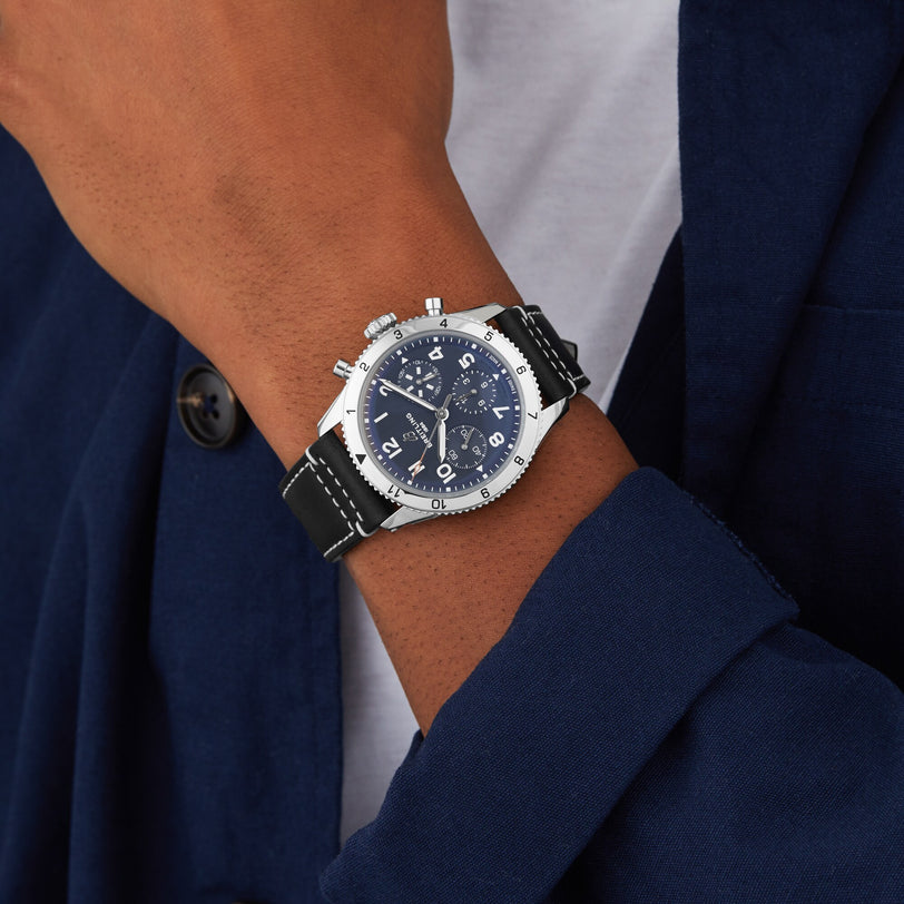 A person in a blue blazer and white shirt showcases the Breitling Classic AVI Chronograph 42 Tribute To Vought F4U, featuring a blue dial, black leather strap, and silver case.