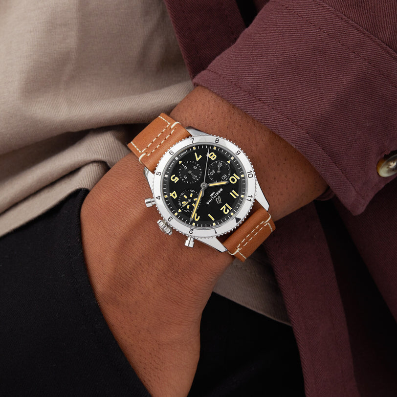 A person wearing a Breitling Classic AVI Chronograph 42 P-51 Mustang with a black dial, tan leather strap, and yellow numerals rests their hand in their pocket.