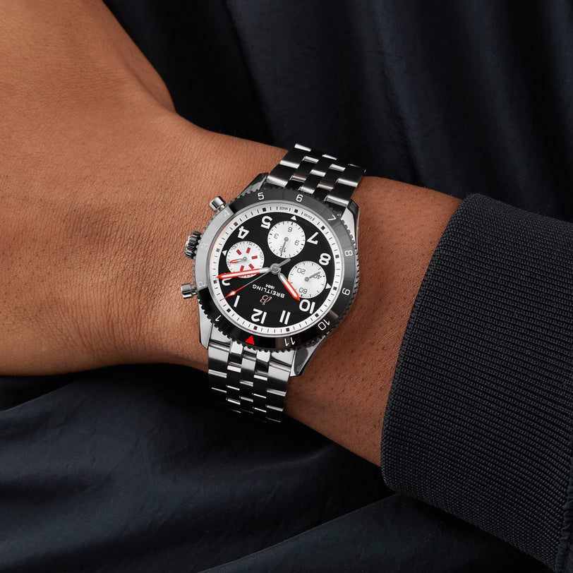 A person wearing a Breitling Classic AVI Chronograph 42 Mosquito, featuring a black and white dial with red accents inspired by aviation history, set against a dark fabric background.