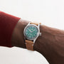 A person wearing the Oris Big Crown X Cervo Volante watch, featuring a green dial with white numerals and a tan leather strap. The Oris brand and classic ProPilot-inspired design are visible on the face.