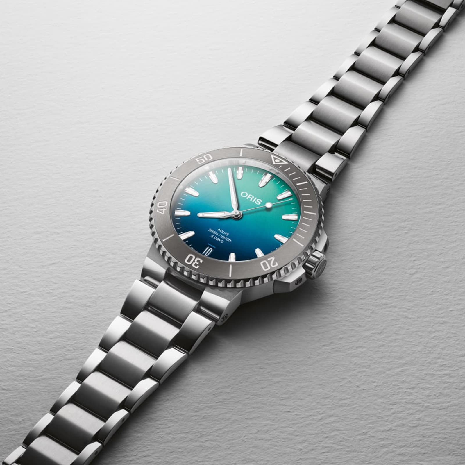 The Oris Great Barrier Reef Limited Edition IV, a stainless steel automatic watch with a blue-green gradient dial and metal bracelet, is displayed on a light gray textured surface.
