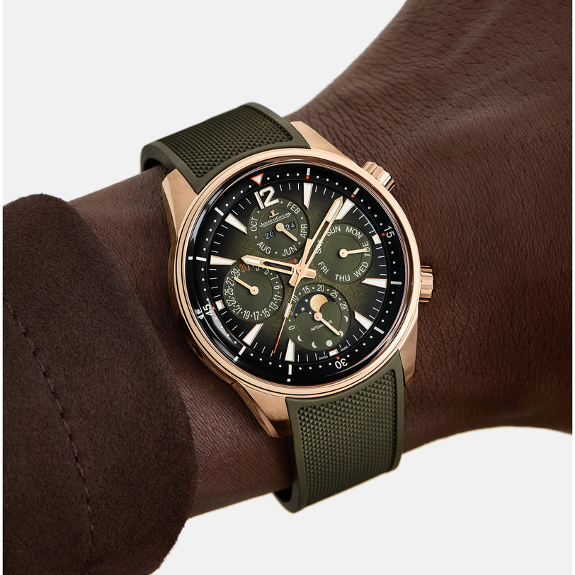A close-up of a wrist with dark brown skin wearing a Jaeger-LeCoultre Polaris Perpetual Calendar gold-tone watch with a green strap, white-marked dial, gold accents, and multiple subdials for day, date, and month; brown sleeve visible.