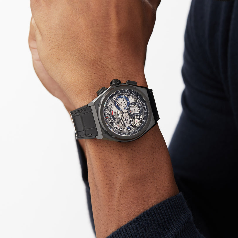 Someone is wearing a Zenith Defy El Primero 21 wristwatch featuring a dark skeleton-style design, black strap, and a visible high-frequency chronograph movement on their left wrist.