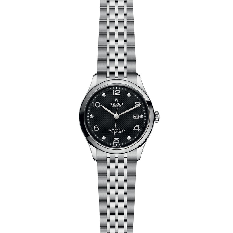 The TUDOR 1926 men’s watch features a silver case, black dial, silver numerals, date at 3 o’clock, and metal link bracelet—reflecting Hans Wilsdorf’s tradition of mechanical timepieces against a white background.
