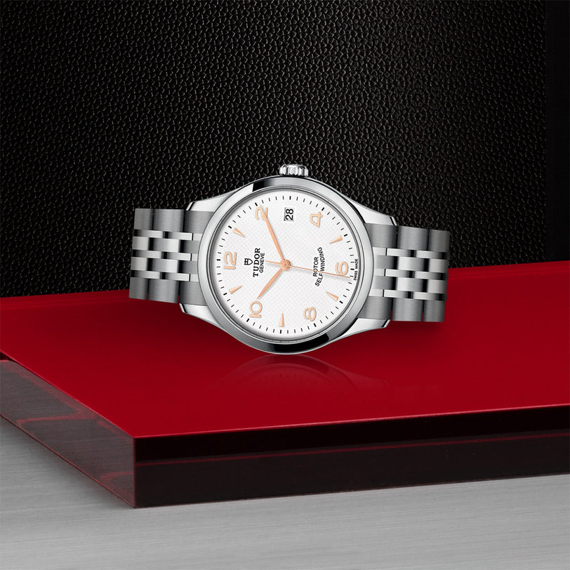 A TUDOR 1926 wristwatch with a silver metal bracelet and white dial is showcased on a glossy red and black surface, set against a textured black background.