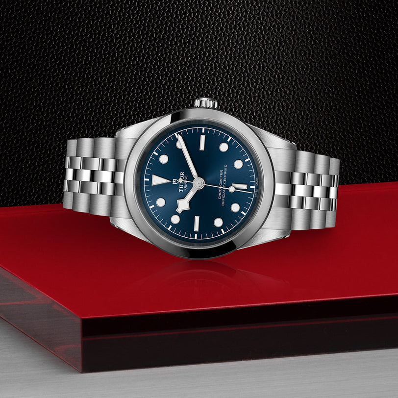 The TUDOR Black Bay One 41, a stainless steel wristwatch with a blue dial and silver bracelet inspired by Black Bay models, is displayed on a red and clear stand against a textured black background.