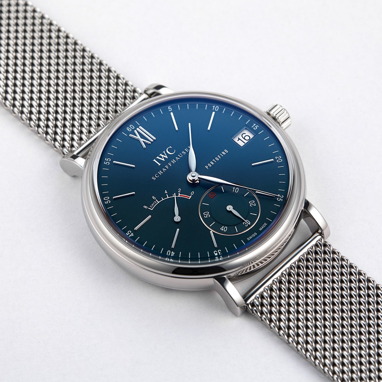 An IWC Schaffhausen Portofino Hand-Wound Eight Days watch with a blue dial, silver mesh band, Roman numeral at 12, and subdials for seconds and date, shown on a white background.