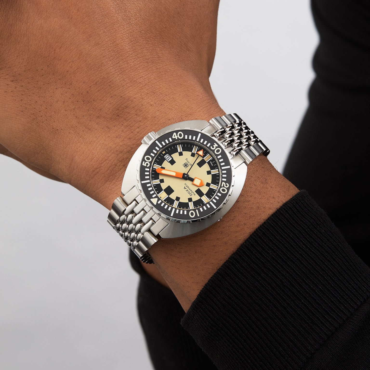 A person wearing a DOXA Army Stainless Steel watch by DOXA, featuring a black and beige dial, orange minute hand, rotating bezel, and stainless steel bracelet.