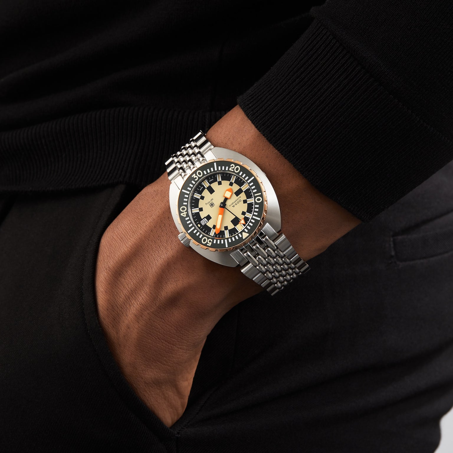 A person wearing a DOXA Army Bronze wristwatch by DOXA, featuring a black and beige dial with Super-LumiNova® luminescent coating and a metal bracelet, hand resting in a pocket.