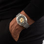 A person wearing a DOXA Army Bronze wristwatch by DOXA, featuring a black and beige dial with Super-LumiNova® luminescent coating and a metal bracelet, hand resting in a pocket.